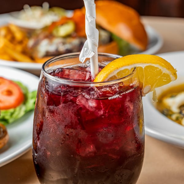 Close-up of a refreshing red drink with ice, a lemon slice, and a straw, with blurred food in the background.