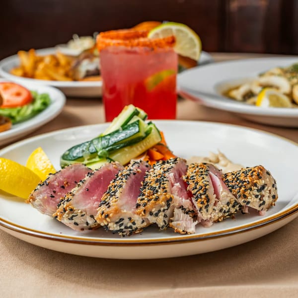 Seared tuna with sesame seeds, lemon wedges, and sliced cucumbers on a white plate.