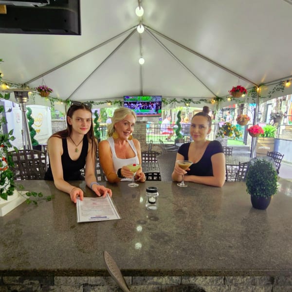 Three women enjoying cocktails at a bar under a white tent at Austin's Ale House.