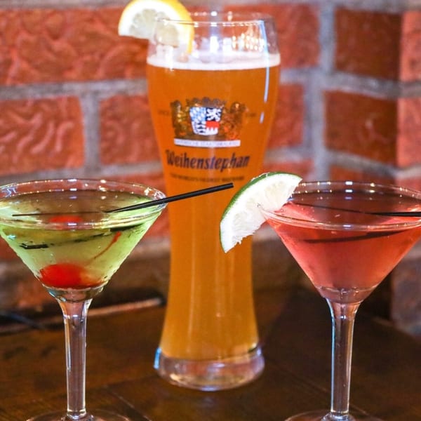 Two colorful martinis and a tall glass of beer with a lemon wedge against a brick wall.