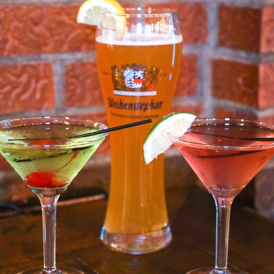 Two colorful martinis and a tall glass of beer with a lemon wedge against a brick wall.