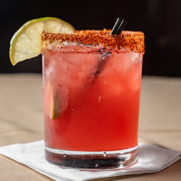 A vibrant red cocktail with a chili-lime rim, garnished with a fresh lime wedge and black straws.